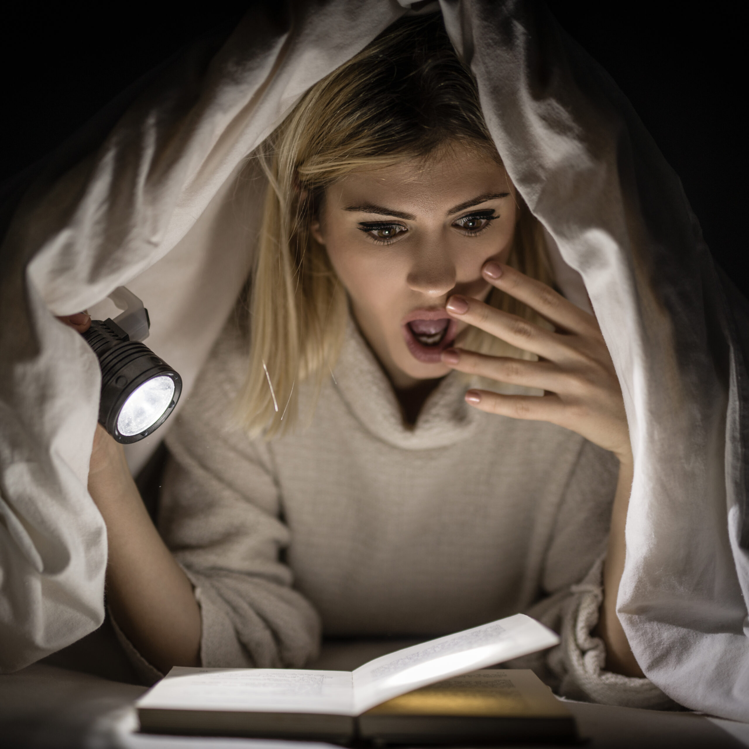 Woman reading a scary story, looking surprised, under a blanket