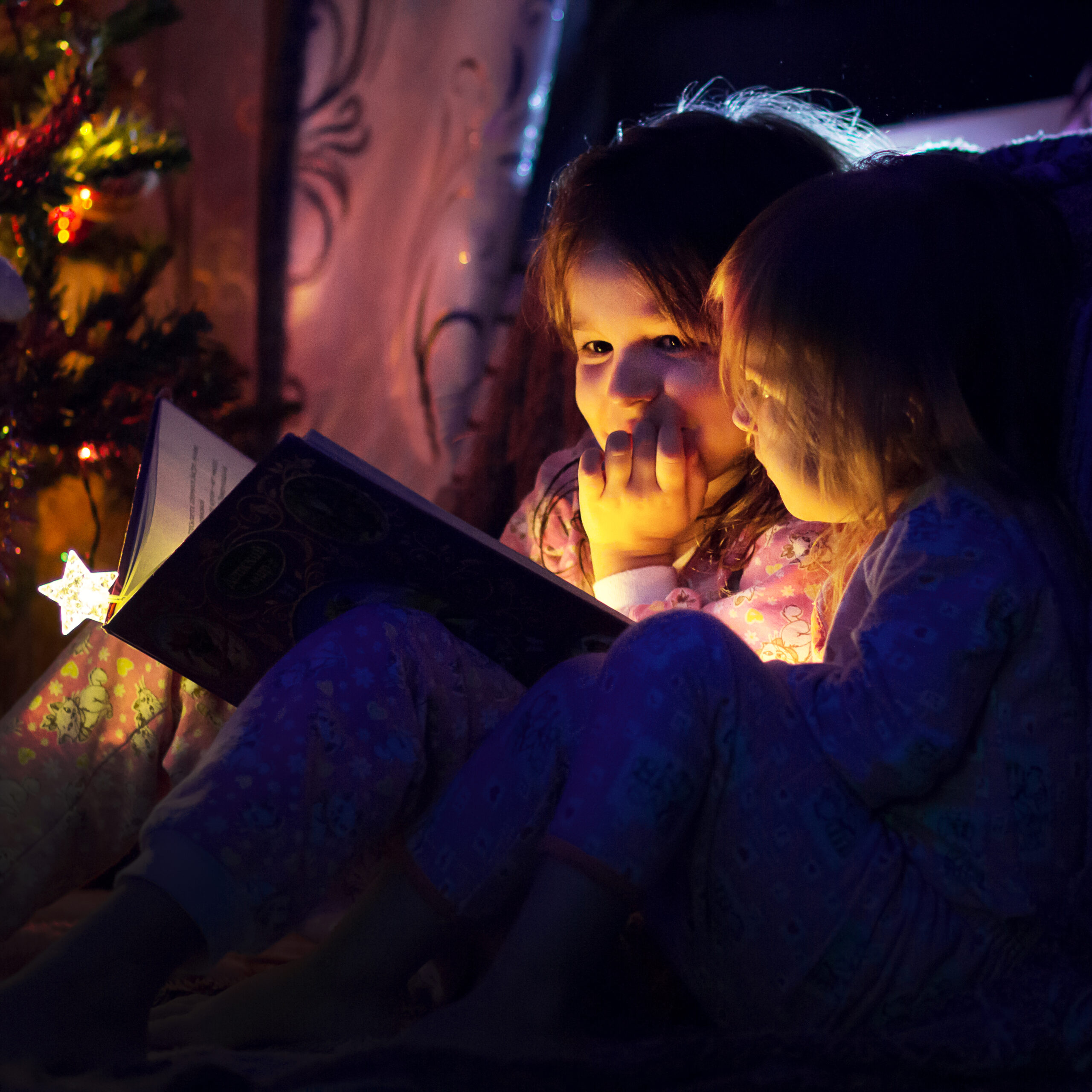 Two sisters read a book by book light near a Christmas tree