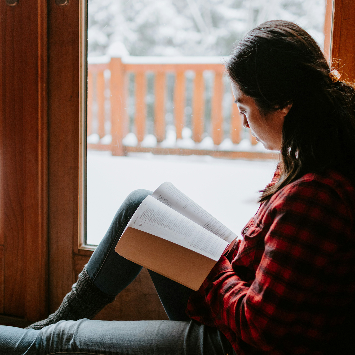 Woman reading next to a window with snow on the ground outside.