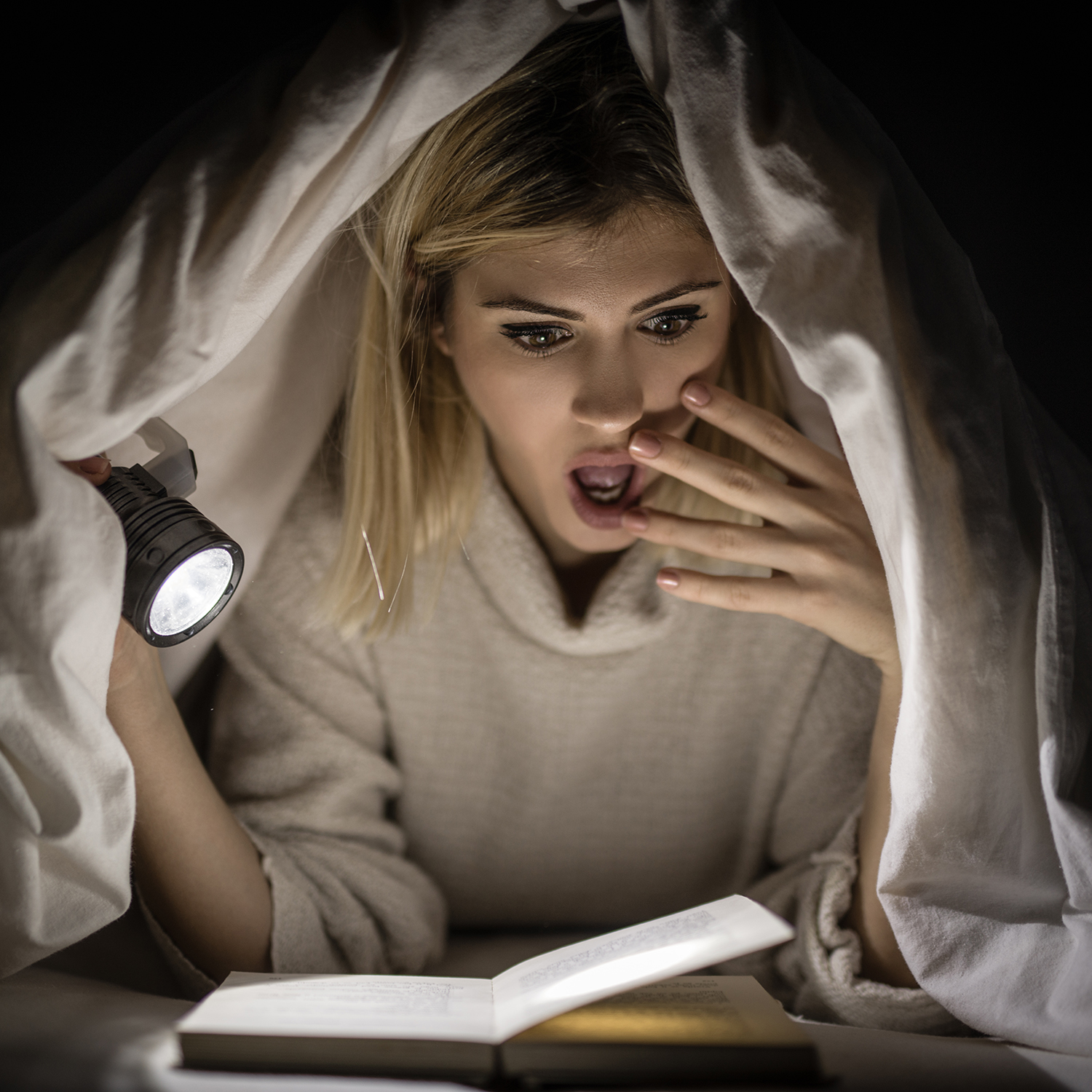 Teen reading a horror book under the covers with a flashlight